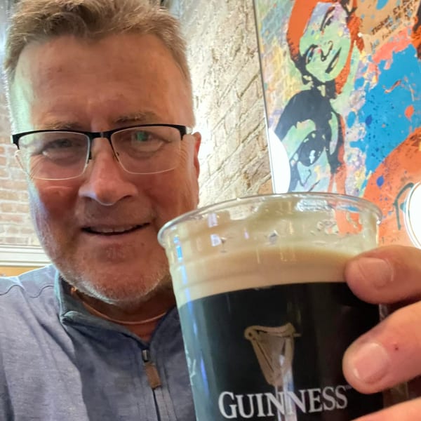 A man in glasses smiles while holding up a pint of Guinness beer at The Harp And Fiddle.