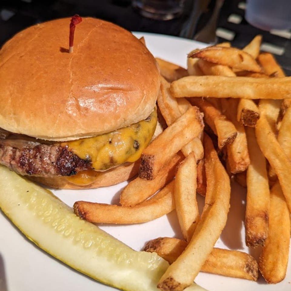 A Harp And Fiddle cheeseburger with fries and a pickle spear on a white plate.