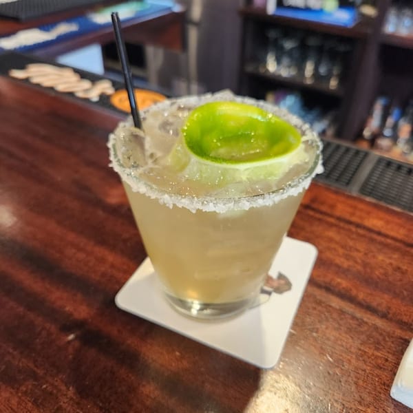 A Harp And Fiddle margarita with a salted rim, lime wedge, and straw on a wooden bar.